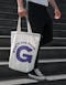 Person carrying the good drop tote bag in white and purple copy saying whatll you have G
